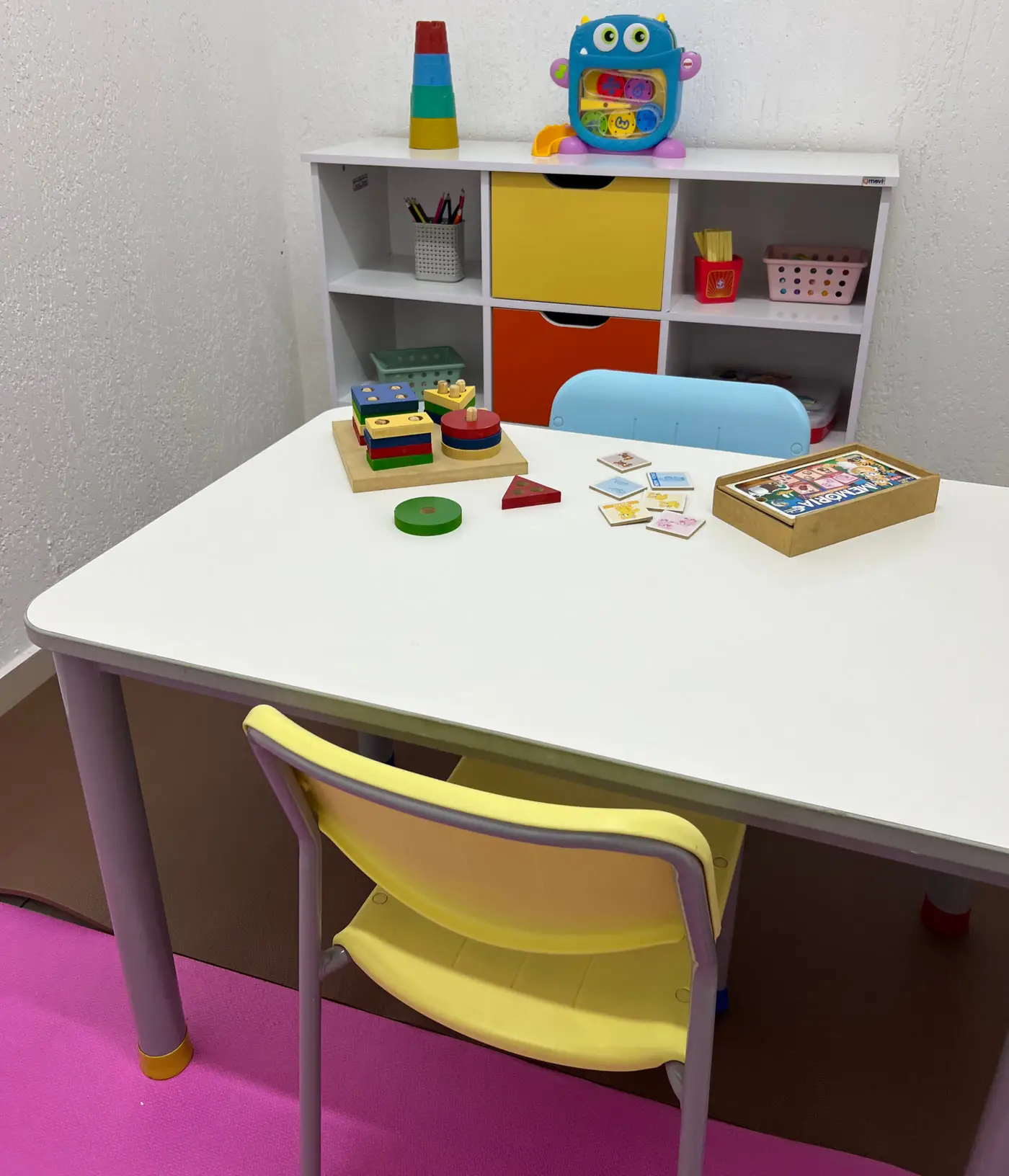 Sala de atendimento com mesa infantil, cadeiras coloridas e brinquedos pedagógicos na estante
