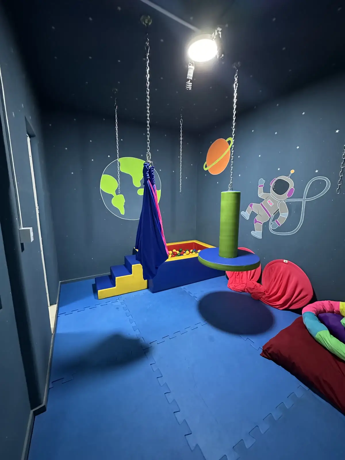 Sala sensorial com piso azul, balanço suspenso, escada de espuma e astronauta pintado na parede