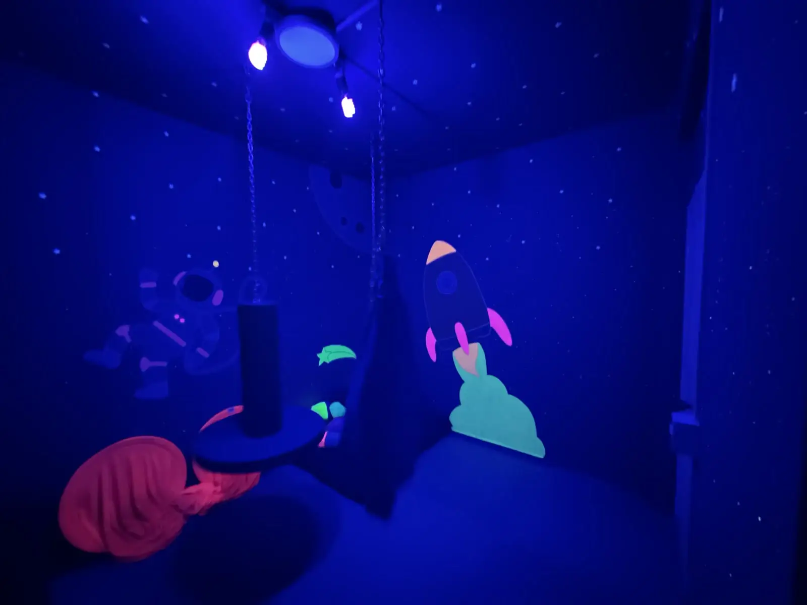 Sala de integração sensorial com luz negra, planetas e foguete pintados nas paredes e equipamentos suspensos